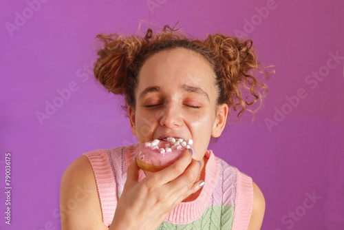 Pretty young caucasian woman eating a donuts with her eyes closed and a candy colored T-shirt on pink background