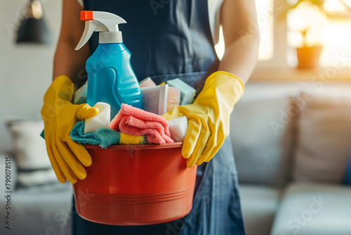 A woman cleaner in an apron and gloves with a red bucket of cleaning products in apartments. Housecleaning with detergents, cleaning supplies. © Oksana Tryndiak