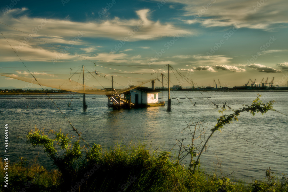 Tranquil waters embrace a picturesque scene of fishing boats and rustic huts along the shores of Pialassa dei Piomboni, near Ravenna