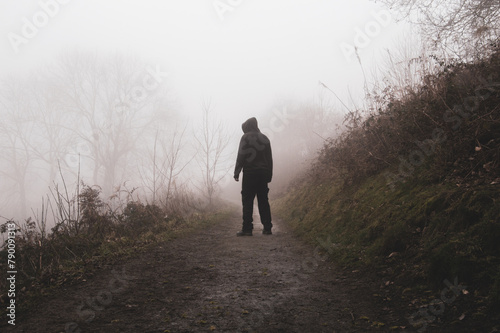 A scary hooded figure. Back to camera. Standing on a path in the countryside. On a spooky foggy winters day