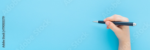 Baby boy hand holding black pen and writing on light blue table background. Pastel color. Closeup. Wide banner. Empty place for text. Top down view.