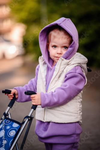 A young girl clad in a violet ensemble attentively maneuvers her toy stroller on an outdoor walk, her gaze reflecting focus and innocence