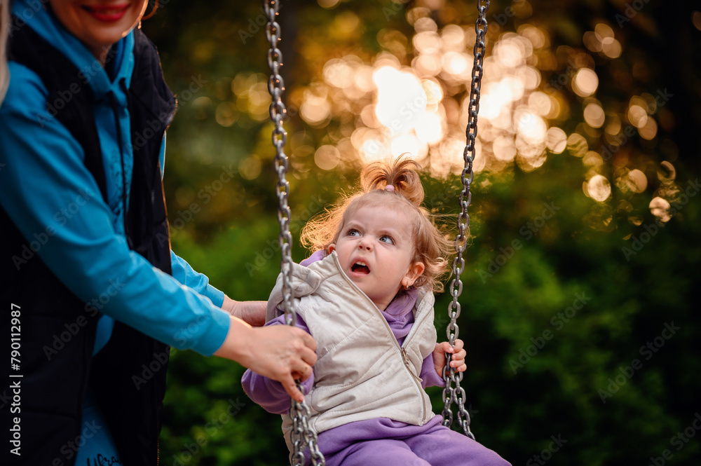 A mother gently supports her little one on a swing, with the toddler's expression of curiosity capturing the simple joys of a new experience A mother gently supports her little one on a swing, with the toddler's expression of curiosity capturing the simple joys of a new experience