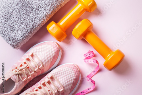 Fitness concept with orange dumbbells, towel,  pink sneakers and measure tape on pink background top view