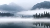 Trees partially obscured by fog on the edge of a calm lake, surrounded by mountains in the distance
