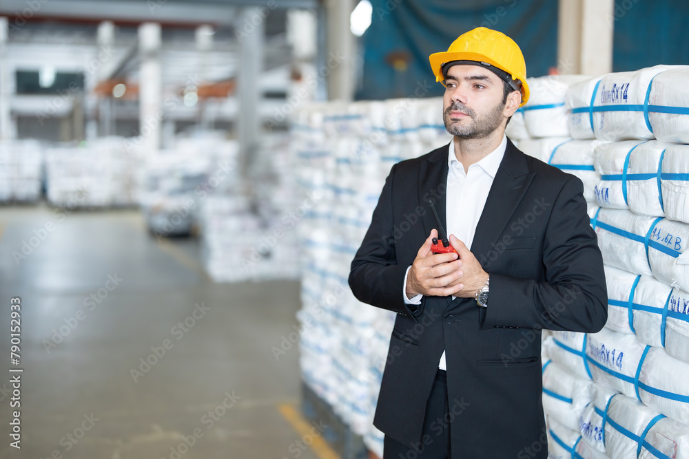 A male European executive engineer stands next to a standard rack of plastic and steel manufacturing plants, wearing a suit and a hard hat, holding a radio, for sale to the international market. A male European executive engineer stands next to a standard rack of plastic and steel manufacturing plants, wearing a suit and a hard hat, holding a radio, for sale to the international market.