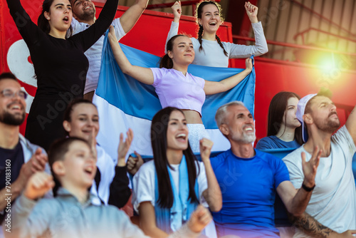 Sport fans cheering at the game on stadium. Wearing blue and white colors to support their team. Celebrating with flags and scarfs.