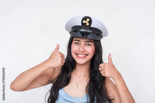 Smiling young Asian woman in captain's hat, making thumbs up sign, isolated on white