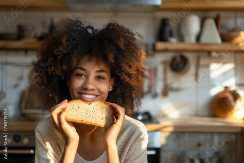 A cheerful young woman with curly hair holding a slice of bread in a sunlit kitchen photo