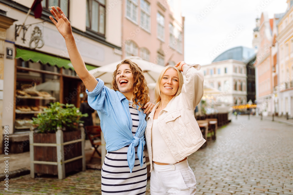 Positive stylish girls smiling on city background.  Female tourists in bright dress enjoys sights of the city on sunny day. Concept of lifestyle, fashion, travel.
