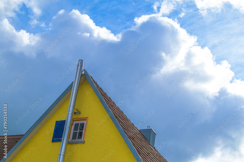 Giebelseite gelbes Haus mit blauen Fensterladen und Edelstahlkamin vor blauem Himmel mit dramatischen Wolken