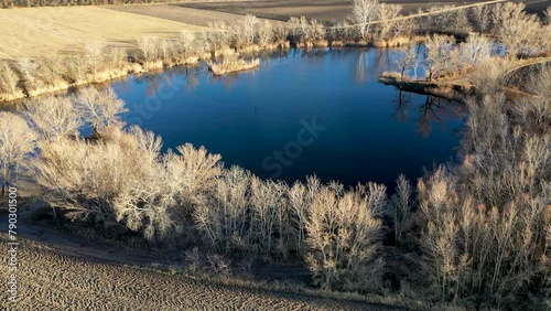 Flying above a small blue lake in the middle of empty fields during winter. Aerial view of the lake and its surrounding trees photo