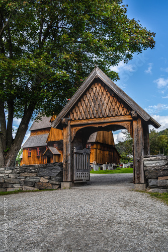 A wooden gate leading to the historic medieval Ringebu Stave Church surrounded by headstones against a summer blue sky with clouds Innlandet county, Norway photo