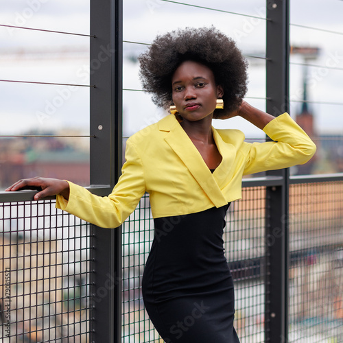 An attractive young African woman is posing on a footbridge with great view over the city. She is wearing a black dress and a beautiful yellow jacket.