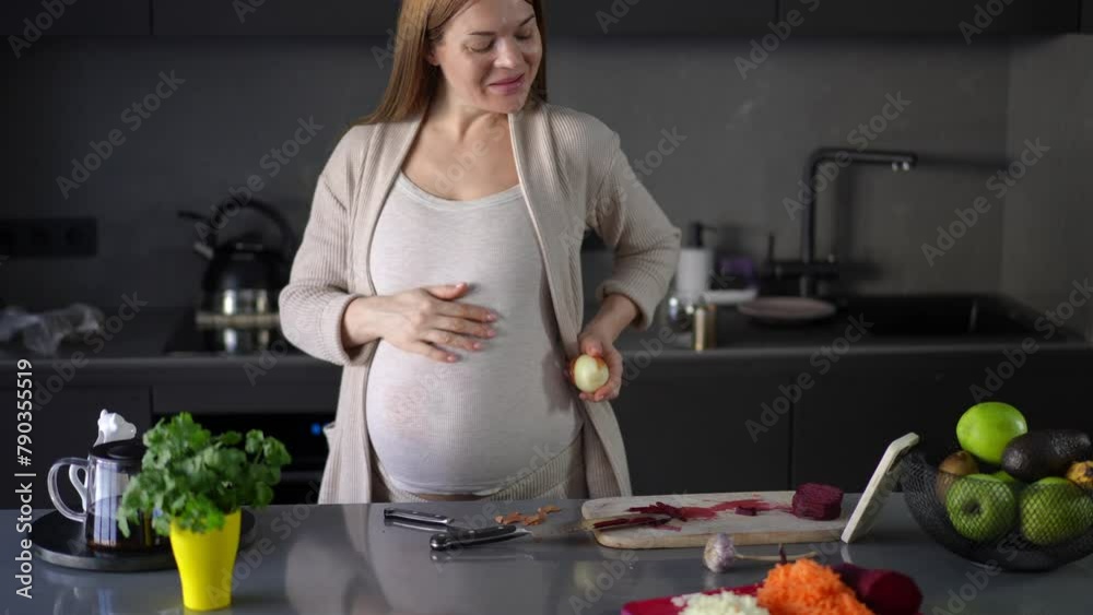 A happy pregnant woman peels onions with a kitchen knife and communicates via video using a mobile phone, standing near the table in the kitchen. A woman talks, smiles and waves her hand at the camera A happy pregnant woman peels onions with a kitchen knife and communicates via video using a mobile phone, standing near the table in the kitchen. A woman talks, smiles and waves her hand at the camera