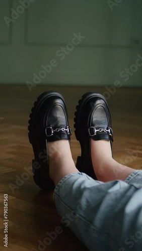 Person wearing black chunky loafers with rolled-up jeans, sitting with feet up on wooden floor. Fashionable footwear concept for design and advertising with copy space.