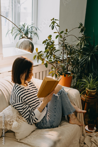 Young brunette woman reading a book on a couch, in soft day light by a window, surrounded by green houseplants.