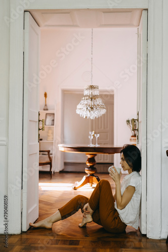 Young brunette woman holding a cup of coffee at home, in a cozy room, sitting on a wooden floor in the doorway.