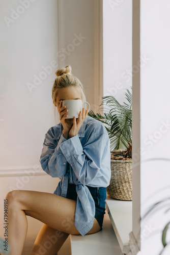 Blonde woman with a cup of coffee at home by the window, sitting on the windowsill, covering face with a white mug.