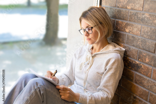 Blonde girl in glasses sits near the window and writes notes in a notebook. Beautiful woman pensively writing with pen in notebook, student, freelancer, cafeteria, sunny day, panoramic window