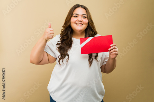 Pretty Hispanic plus-size woman in a photo studio holding a red gift card and giving a thumbs up sign