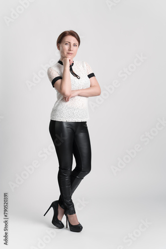 Business woman wearing white short sleeve blouse, black leather tight trousers and high heeled shoes looking at camera with one hand raised to chin, standing legs crossed at ankle on white background