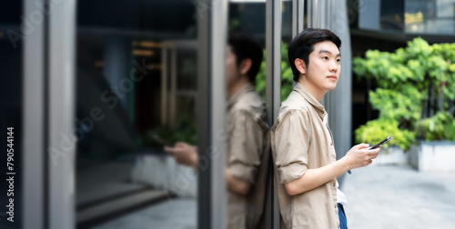 Portrait of handsome Asian student using smartphone. A young man standing outdoor happy smiling with holding mobile phone