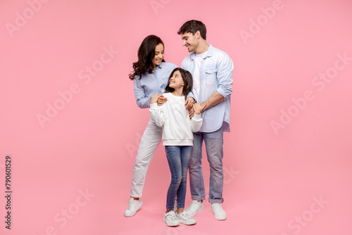 European parents embracing their happy daughter looking at pretty girl, standing together on pink studio background, full length shot