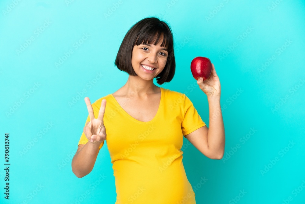 Pregnant woman holding an apple isolated on blue background smiling and showing victory sign Pregnant woman holding an apple isolated on blue background smiling and showing victory sign