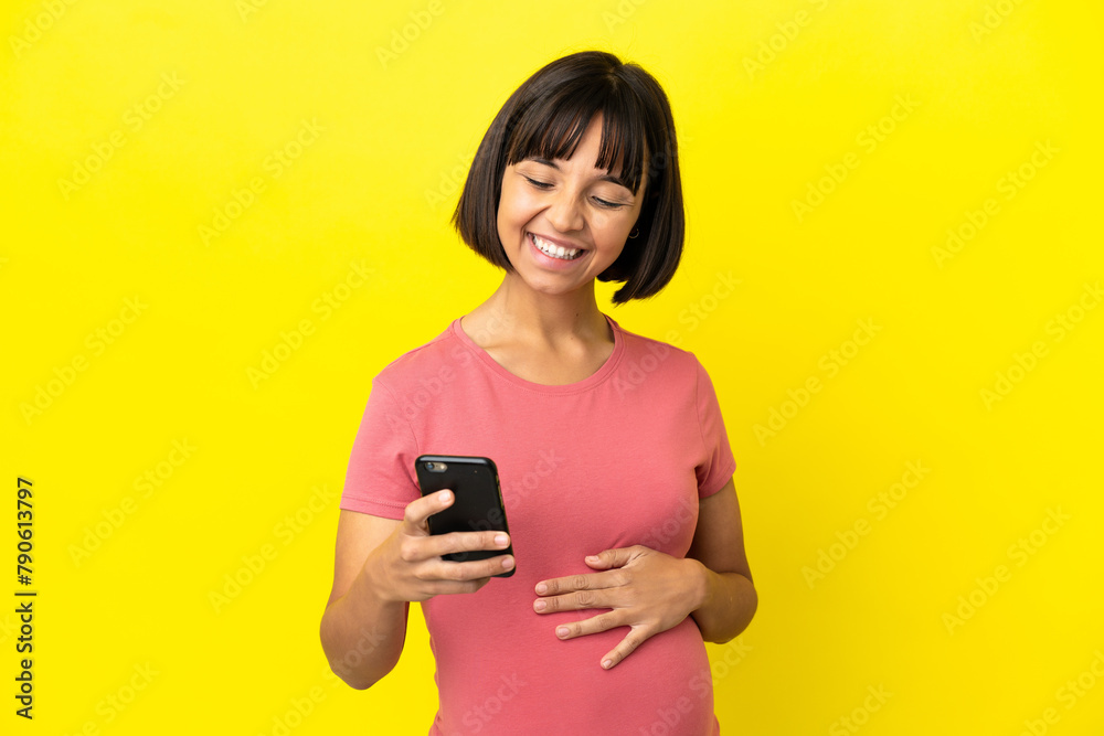 Young mixed race pregnant woman isolated on yellow background sending a message or email with the mobile Young mixed race pregnant woman isolated on yellow background sending a message or email with the mobile