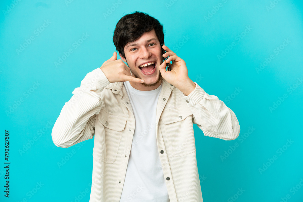 Young Russian man using mobile phone isolated on blue background making phone gesture. Call me back sign Young Russian man using mobile phone isolated on blue background making phone gesture. Call me back sign