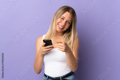 Young blonde woman using mobile phone isolated on purple background points finger at you
