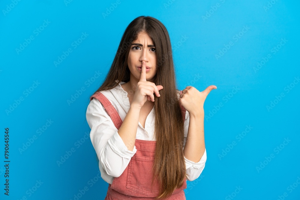 Young caucasian woman isolated on blue background pointing to the side and doing silence gesture Young caucasian woman isolated on blue background pointing to the side and doing silence gesture