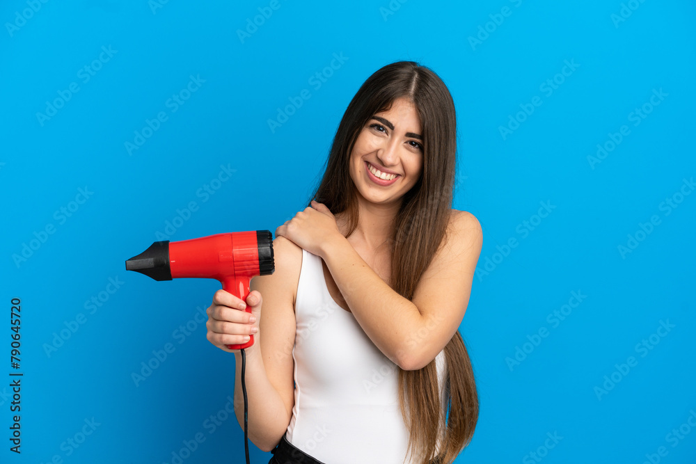 Young caucasian woman holding a hairdryer isolated on blue background laughing Young caucasian woman holding a hairdryer isolated on blue background laughing