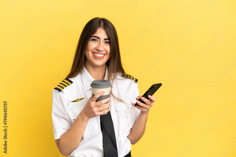 Airplane pilot isolated on yellow background holding coffee to take away and a mobile Airplane pilot isolated on yellow background holding coffee to take away and a mobile