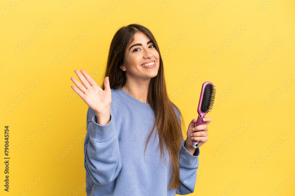 Young caucasian woman holding hairbrush isolated on blue background saluting with hand with happy expression Young caucasian woman holding hairbrush isolated on blue background saluting with hand with happy expression