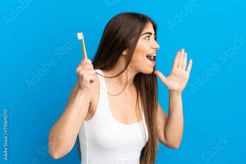 Young caucasian woman brushing teeth isolated on blue background shouting with mouth wide open to the side