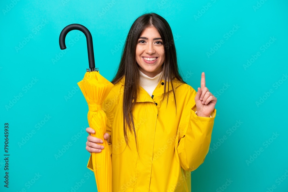 Young caucasian woman with rainproof coat and umbrella isolated on blue background pointing up a great idea Young caucasian woman with rainproof coat and umbrella isolated on blue background pointing up a great idea