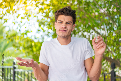 Young caucasian man holding invisible braces at outdoors making doubts gesture while lifting the shoulders