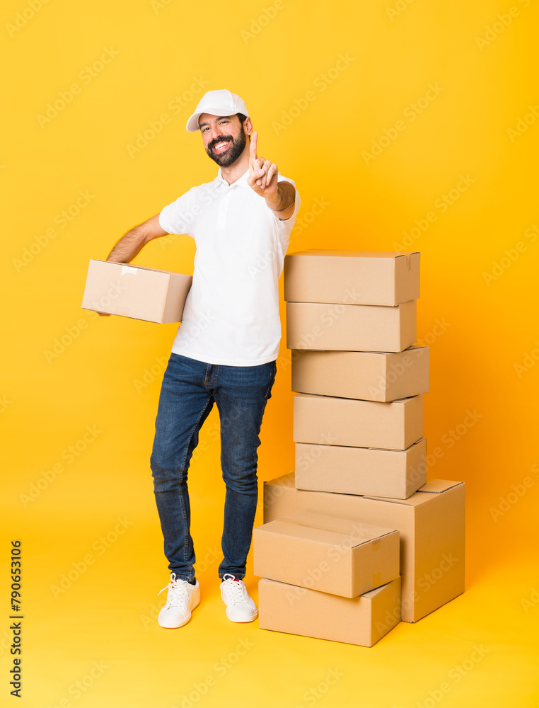 Full-length shot of delivery man among boxes over isolated yellow background showing and lifting a finger Full-length shot of delivery man among boxes over isolated yellow background showing and lifting a finger