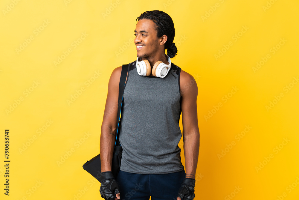 Young sport African American man with braids with bag isolated on yellow background looking to the side and smiling Young sport African American man with braids with bag isolated on yellow background looking to the side and smiling
