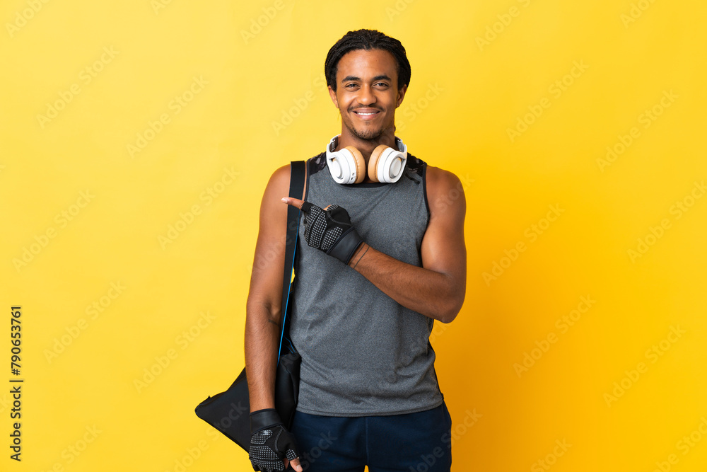 Young sport African American man with braids with bag isolated on yellow background pointing to the side to present a product Young sport African American man with braids with bag isolated on yellow background pointing to the side to present a product
