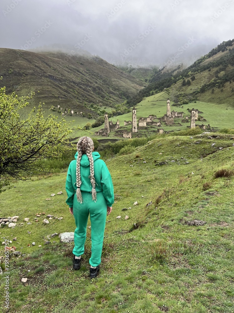 A girl on the background of the medieval Targim tower complex in the Caucasus mountains, surrounded by greenery. The Caucasus Mountains on a cloudy day in Ingushetia. Russia