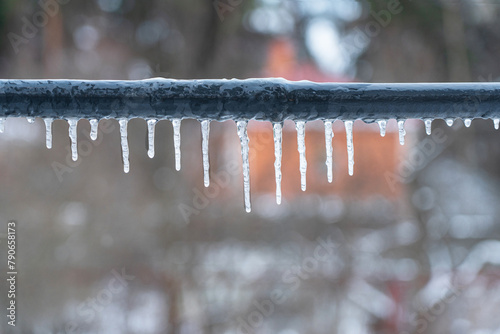 Several icicles of different lengths hang from the gray pipe of the crossbar. A blurry rural landscape is visible in the background. Background.
