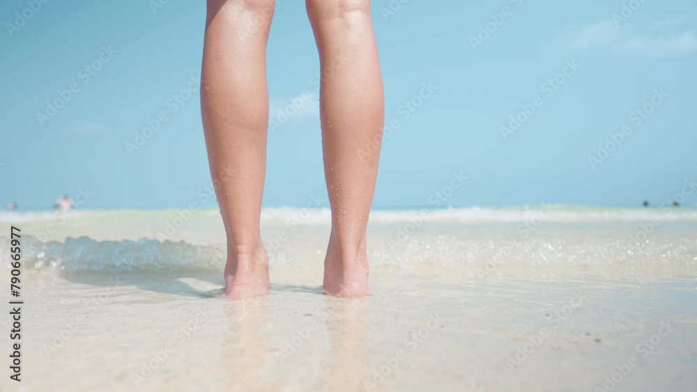Low angle shot of female smooth skin legs standing and soaking in sea water with blue sky on background, ocean waves hitting feet on white sand beach, summer islands vacation, barefoot on the beach Low angle shot of female smooth skin legs standing and soaking in sea water with blue sky on background, ocean waves hitting feet on white sand beach, summer islands vacation, barefoot on the beach