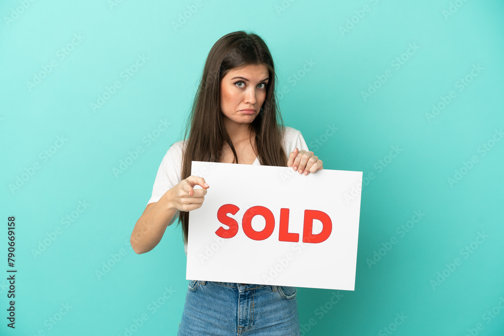 Young caucasian woman isolated on blue background holding a placard with text SOLD and pointing to the front Young caucasian woman isolated on blue background holding a placard with text SOLD and pointing to the front