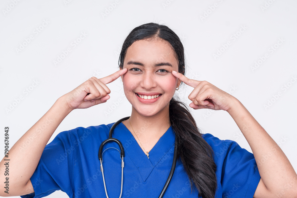 Smiling Asian female nurse in blue scrub suit pointing to eyes, isolated on white Smiling Asian female nurse in blue scrub suit pointing to eyes, isolated on white
