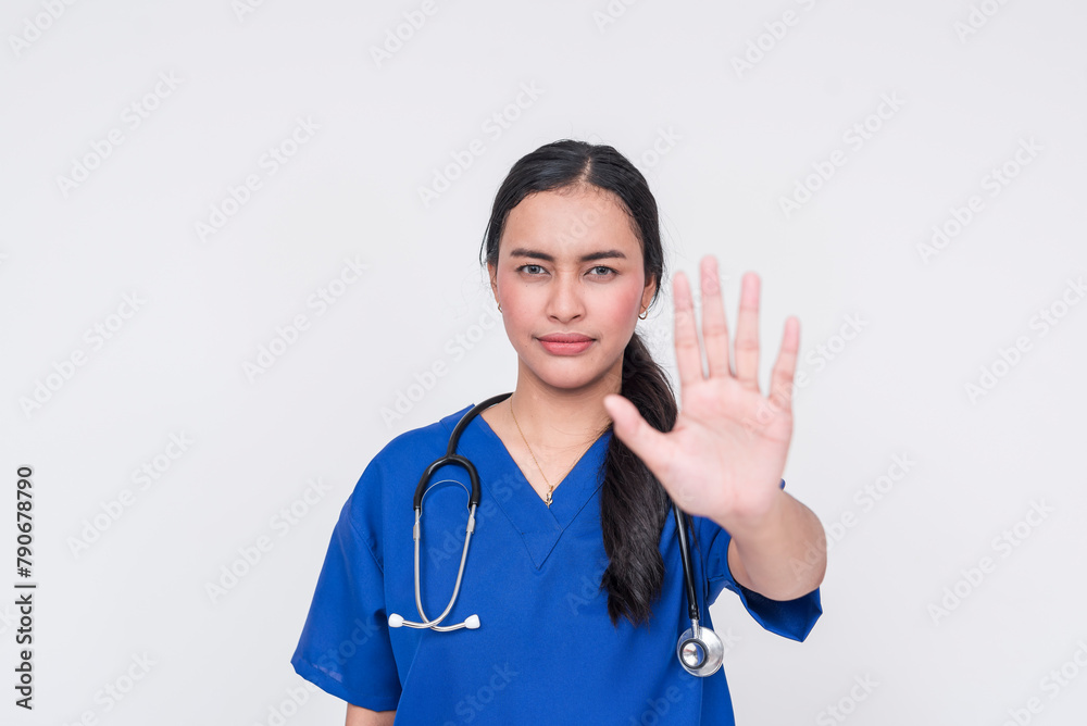 A young female nurse wearing a blue scrub suit gestures to stop, symbolizing caution or restriction, isolated on a white background. A young female nurse wearing a blue scrub suit gestures to stop, symbolizing caution or restriction, isolated on a white background.