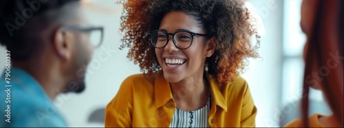 Closeup of two people talking and smiling during an interview, one person is African American woman with curly hair wearing glasses in yellow jacket sitting at the table, another man behind her 