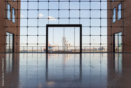 Looking through a vast, contemporary glass window, a prominent glass door sits centrally, flanked by building structures on either side. In the background, silhouettes of harbor cranes and industrial 